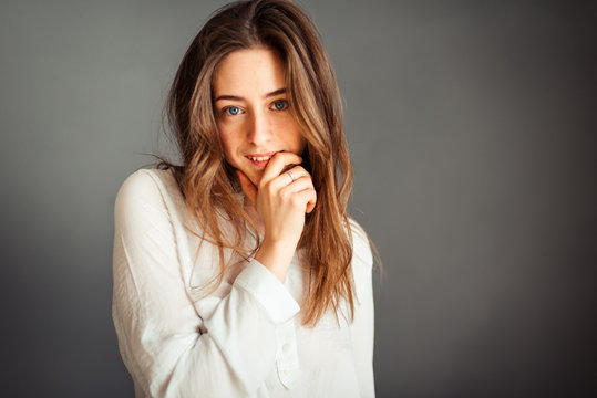 Young smiling girl in a white shirt hand near the mouth, the second in the pocket, tousled hair, isolator on a gray background. No retouching. Without makeup.