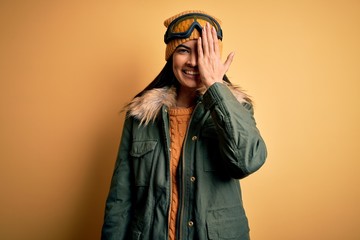 Young beautiful hispanic woman wearing ski glasses and coat for winter weather covering one eye with hand, confident smile on face and surprise emotion.