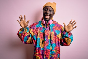 Young handsome african american man wearing colorful coat and cap over pink background showing and pointing up with fingers number ten while smiling confident and happy.
