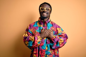 Handsome african american man wearing colorful coat and glasses over yellow background smiling with hands on chest with closed eyes and grateful gesture on face. Health concept.