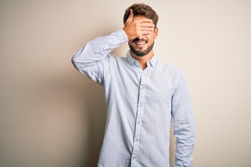 Obraz premium Young handsome man with beard wearing striped shirt standing over white background smiling and laughing with hand on face covering eyes for surprise. Blind concept.