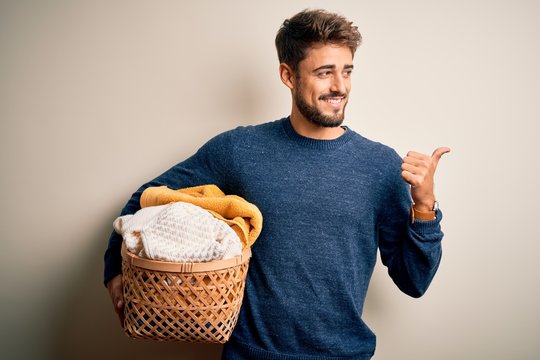 Young man doing housework holding wicker basket with clothes over white background pointing and showing with thumb up to the side with happy face smiling