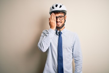 Young businessman wearing glasses and bike helmet standing over isolated white bakground covering one eye with hand, confident smile on face and surprise emotion.