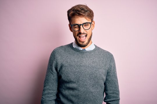 Young handsome man with beard wearing glasses and sweater standing over pink background winking looking at the camera with sexy expression, cheerful and happy face.
