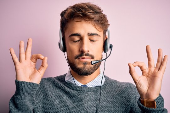 Young call center agent man with beard wearing headset over isolated pink background relax and smiling with eyes closed doing meditation gesture with fingers. Yoga concept.