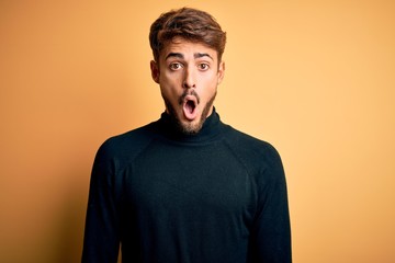 Obraz premium Young handsome man with beard wearing turtleneck sweater standing over yellow background afraid and shocked with surprise expression, fear and excited face.