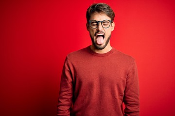 Young handsome man with beard wearing glasses and sweater standing over red background sticking tongue out happy with funny expression. Emotion concept.