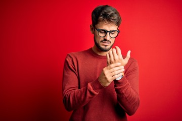 Young handsome man with beard wearing glasses and sweater standing over red background Suffering...