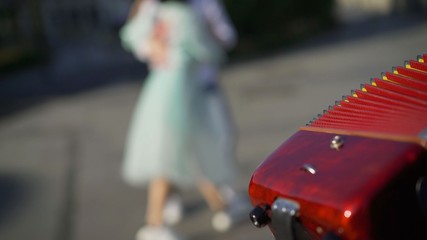 An accordionist is playing outside, and a young couple is dancing. The accordion is in focus, and the dancing couple is out of focus.
