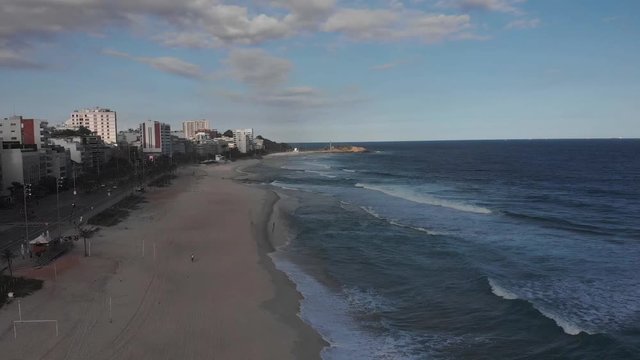 Aerial descend showing an almost empty Ipanema beach in late afternoon with the Arpoador rock in the background against a blue sky with clouds at sunset during the COVID-19 Corona virus outbreak