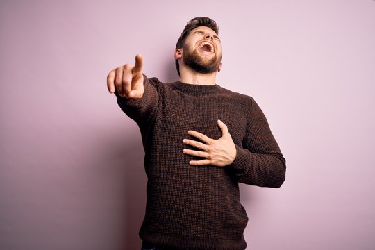 Young blond man with beard and blue eyes wearing casual sweater over pink background laughing at you, pointing finger to the camera with hand over body, shame expression