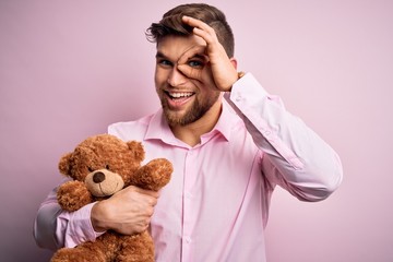 Young blond man with beard and blue eyes holding teddy bear toy over pink background with happy...