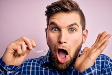 Young blond man with beard and blue eyes holding dental aligner over pink background very happy and excited, winner expression celebrating victory screaming with big smile and raised hands