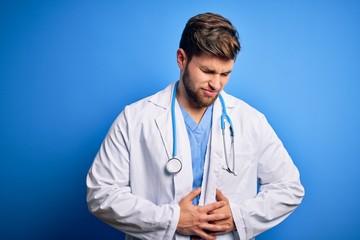 Young blond doctor man with beard and blue eyes wearing white coat and stethoscope with hand on stomach because indigestion, painful illness feeling unwell. Ache concept.