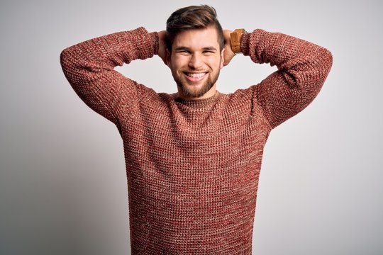 Young blond man with beard and blue eyes wearing casual sweater over white background relaxing and stretching, arms and hands behind head and neck smiling happy
