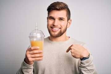 Young blond man with beard and blue eyes drinking healthy orange smoothie with surprise face pointing finger to himself