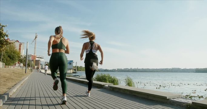 Tracking shot of anonymous female athletes running on paved path near sea water during fitness workout on sunny day