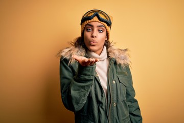 Young african american afro skier girl wearing snow sportswear and ski goggles looking at the camera blowing a kiss with hand on air being lovely and sexy. Love expression.