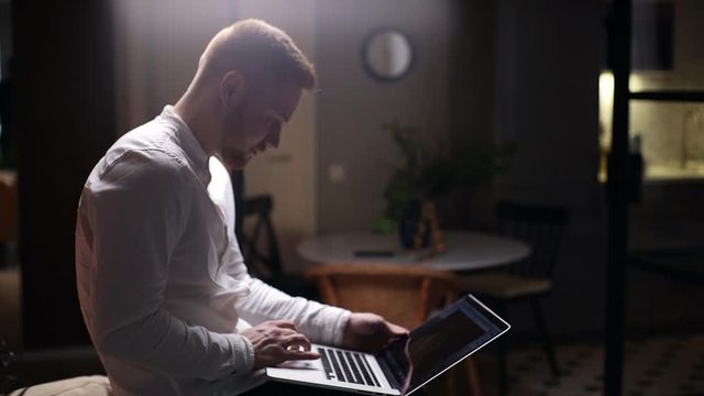 Confident handsome man using laptop while sitting in dark living room, remote working from home. Concept of remote working in home during Coronavirus COVID-19 Pandemic. Shooting in slow motion.