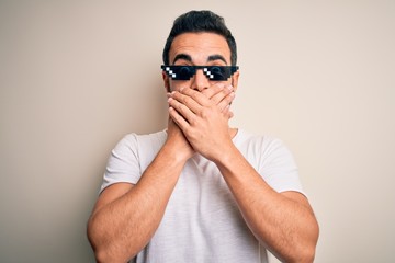 Obraz premium Young handsome man with beard wearing funny thug life sunglasses over white background shocked covering mouth with hands for mistake. Secret concept.