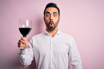 Young handsome man with beard drinking glass of red wine over isolated pink background scared in...