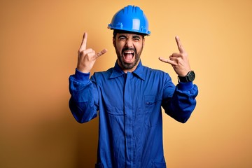 Mechanic man with beard wearing blue uniform and safety helmet over yellow background shouting with...