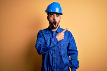 Mechanic man with beard wearing blue uniform and safety helmet over yellow background Surprised...