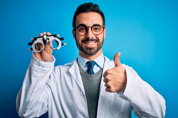 Young handsome optical man with beard holding optometry glasses over blue background doing happy thumbs up gesture with hand. Approving expression looking at the camera showing success.