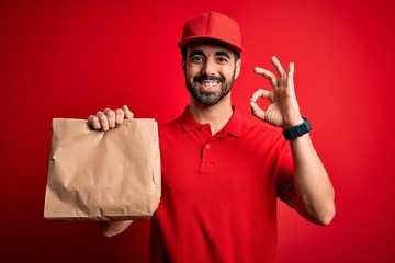 Young handsome delivery man with beard wearing cap holding takeaway paper bag with food doing ok sign with fingers, excellent symbol