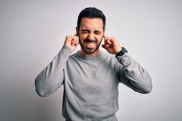 Young handsome man with beard wearing casual sweater standing over white background covering ears with fingers with annoyed expression for the noise of loud music. Deaf concept.