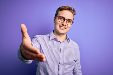 Naklejka premium Young handsome redhead man wearing casual shirt and glasses over purple background smiling friendly offering handshake as greeting and welcoming. Successful business.