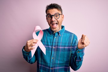 Young handsome man holding pink cancer ribbon symbol over isolated background screaming proud and celebrating victory and success very excited, cheering emotion