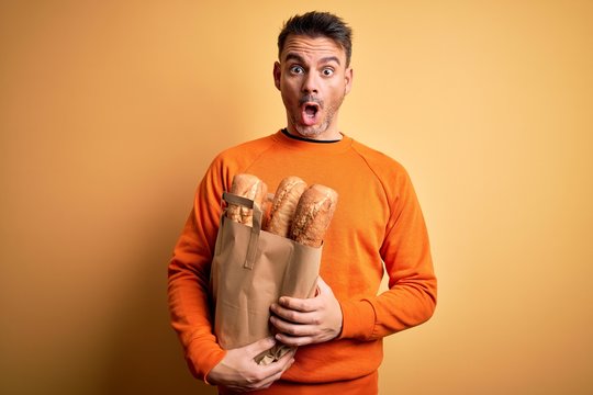 Young handsome man holding paper bag with bread over isolated yellow background scared in shock with a surprise face, afraid and excited with fear expression
