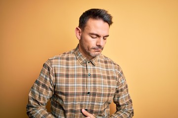 Young handsome man wearing casual shirt standing over isolated yellow background with hand on stomach because indigestion, painful illness feeling unwell. Ache concept.