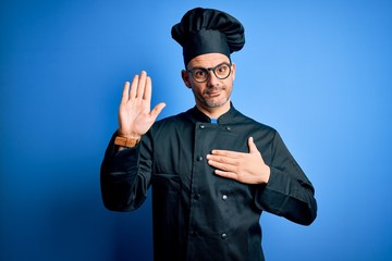 Young handsome chef man wearing cooker uniform and hat over isolated blue background Swearing with...
