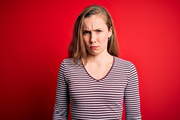Young beautiful blonde woman wearing casual striped t-shirt over isolated red background skeptic...