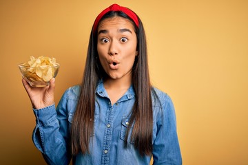 Young beautiful asian woman holding bowl with chips potatoes over isolated yellow background scared in shock with a surprise face, afraid and excited with fear expression