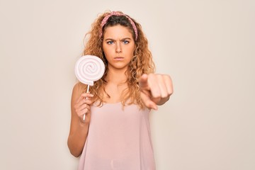 Young beautiful blonde woman with blue eyes eating sweet candy lollipop over pink background pointing with finger to the camera and to you, hand sign, positive and confident gesture from the front