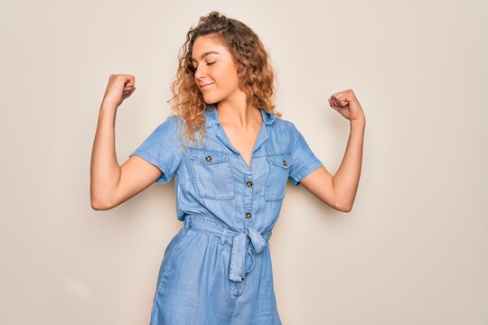 Young beautiful woman with blue eyes wearing casual denim dress over white background showing arms muscles smiling proud. Fitness concept.