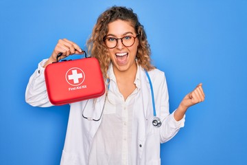 Young beautiful blonde doctor woman with blue eyes wearing stethoscope holding first aid kit...