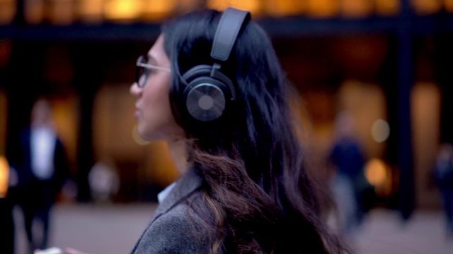 South Asian manager in spectacles and electronic equipment walking at city street in metropolitan downtown and listening knowledge audio book, Beautiful professional in bluetooth technology outdoors
