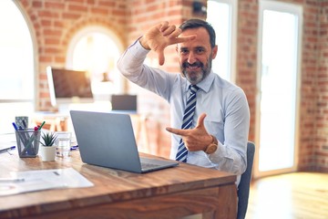 Middle age handsome businessman wearing tie sitting using laptop at the office smiling making frame with hands and fingers with happy face. Creativity and photography concept.