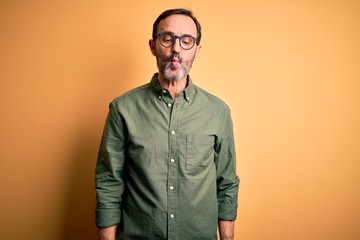 Fototapeta premium Middle age hoary man wearing casual green shirt and glasses over isolated yellow background making fish face with lips, crazy and comical gesture. Funny expression.