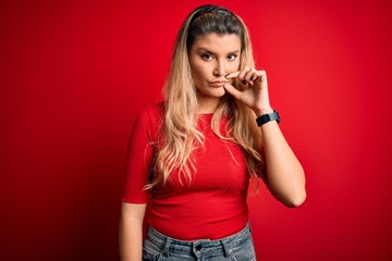 Young beautiful blonde woman wearing casual t-shirt standing over isolated red background mouth and...