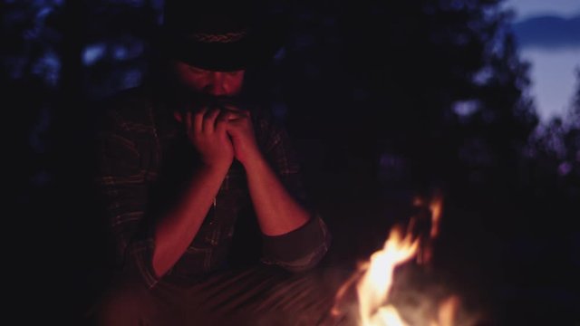 Cowboy plays the harmonica by a campfire at night in slow motion