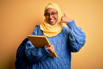 Young African American student woman wearing muslim hijab and backpack holding book smiling doing phone gesture with hand and fingers like talking on the telephone. Communicating concepts.