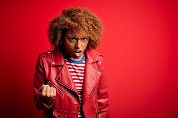 Young beautiful African American afro woman with curly hair wearing casual red jacket angry and mad raising fist frustrated and furious while shouting with anger. Rage and aggressive concept.