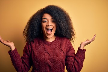 Young beautiful african american woman with afro hair standing over yellow isolated background celebrating crazy and amazed for success with arms raised and open eyes screaming excited. Winner concept