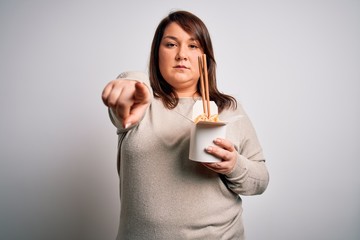 Beautiful plus size woman eating asian noodles food from box over isolated background pointing with finger to the camera and to you, hand sign, positive and confident gesture from the front