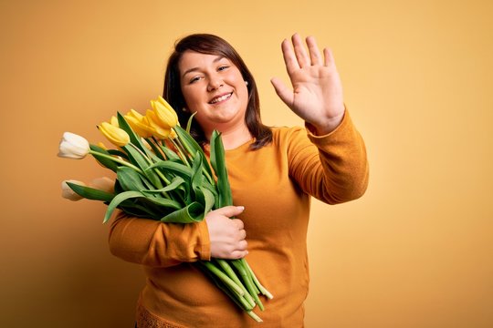 Beautiful plus size woman holding romantic bouquet of natural tulips flowers over yellow background Waiving saying hello happy and smiling, friendly welcome gesture